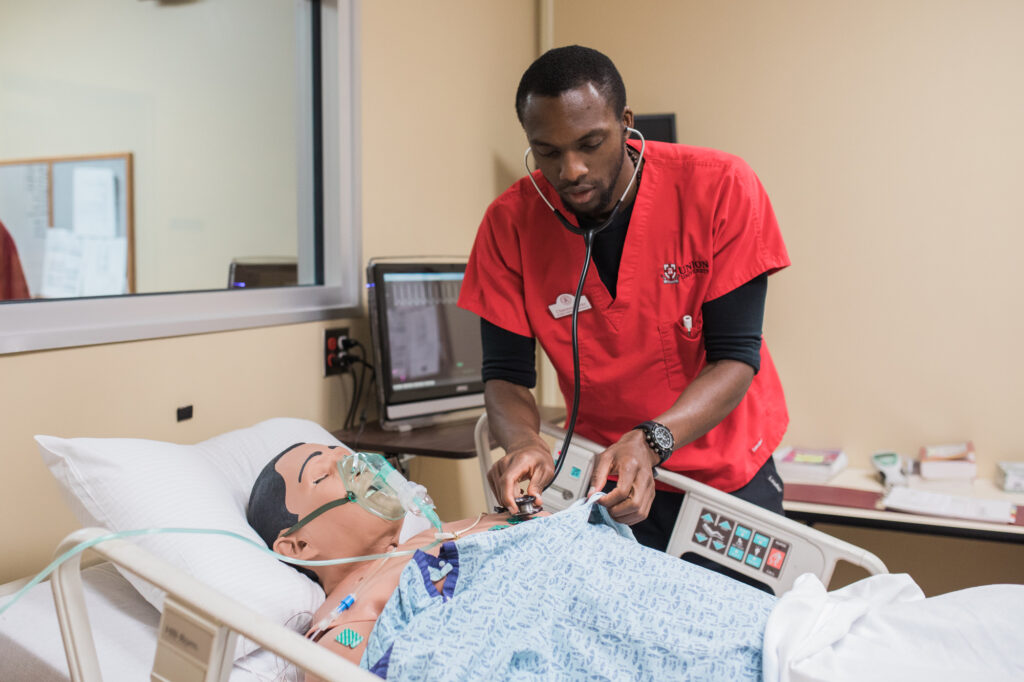 A person in red scrubs uses a stethoscope on a medical mannequin in a hospital bed. The mannequin has an oxygen mask, and there are monitors and medical equipment in the room.