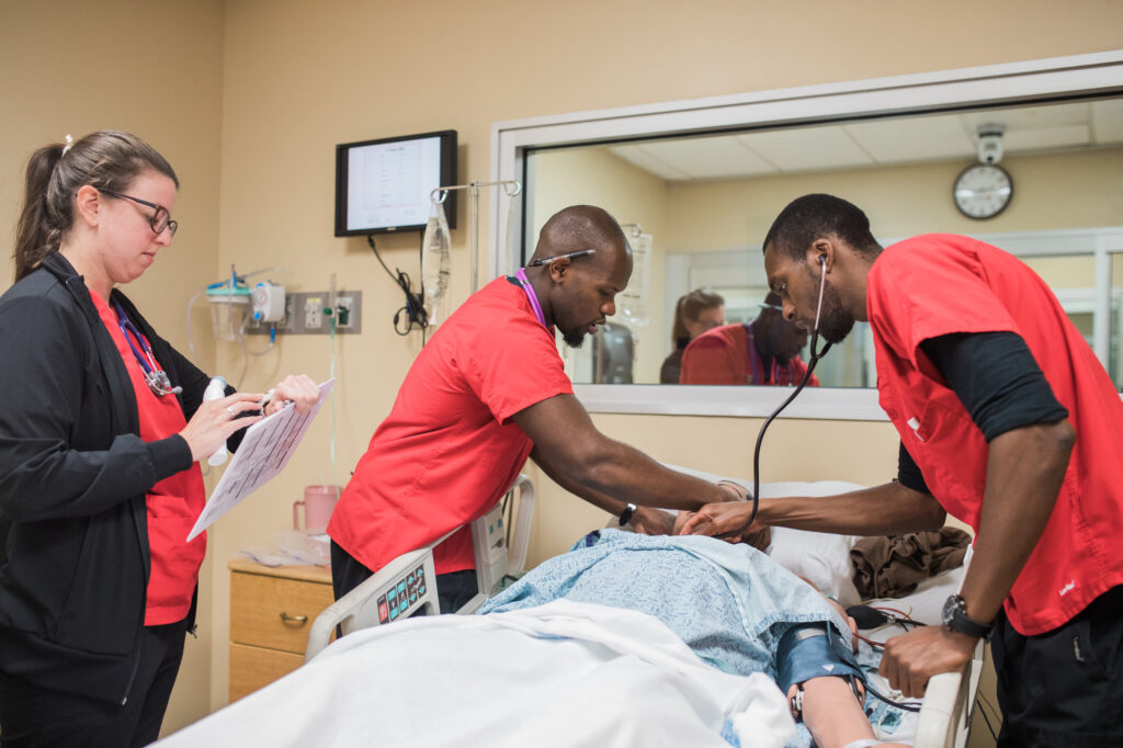 Three healthcare professionals in red uniforms and one in black attend to a patient lying in a hospital bed. Two are checking the patient, while one reviews a chart. Medical equipment and a clock are visible in the room.