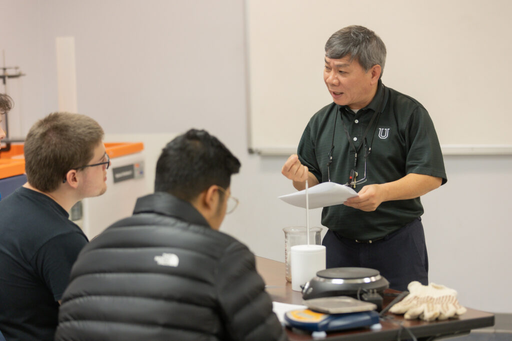 A teacher stands beside a table with lab equipment, holding papers and speaking to two students who are listening attentively. The students are seated and facing the teacher. A whiteboard is visible in the background.