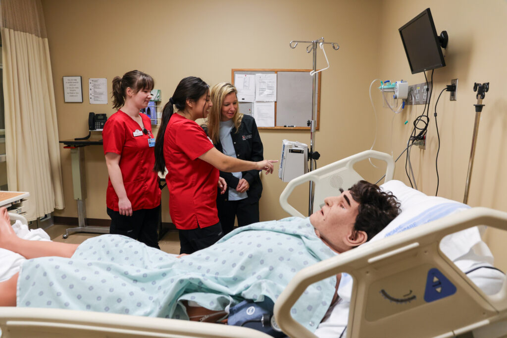 Three healthcare workers stand by a hospital bed, observing a medical mannequin in a patient gown. The room has medical equipment, a monitor on the wall, and a dry erase board. The setting appears to be a training simulation.