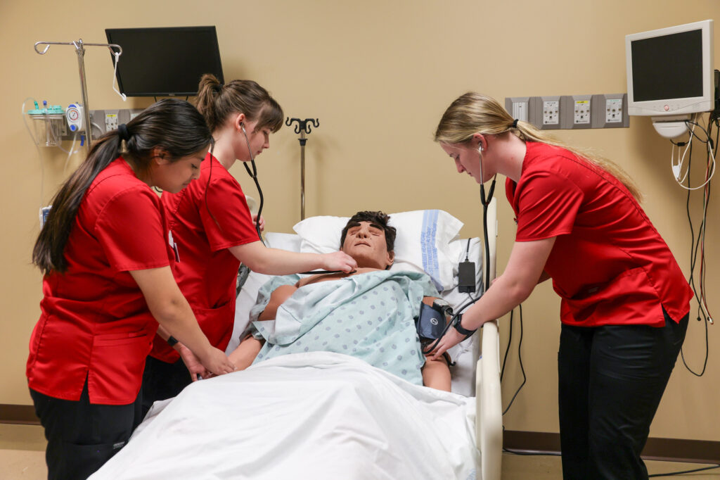 Three healthcare workers in red scrubs check the vital signs of a patient lying in a hospital bed, using a stethoscope and blood pressure cuff, in a medical room with monitors and equipment.