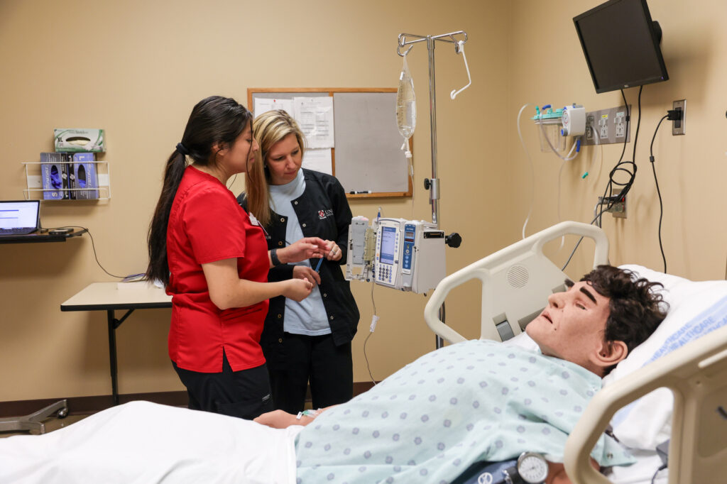 Two medical professionals stand beside a hospital bed with a patient mannequin. Medical equipment, monitors, and IV stand are visible in the room. The professionals are focused on their task.