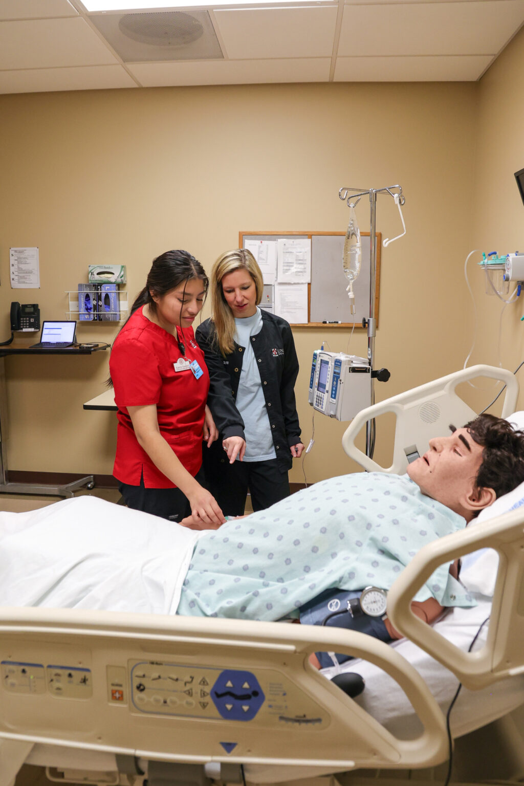 Two medical professionals stand beside a hospital bed, interacting with a medical mannequin patient in a simulation room. Medical equipment and a computer are visible in the background.