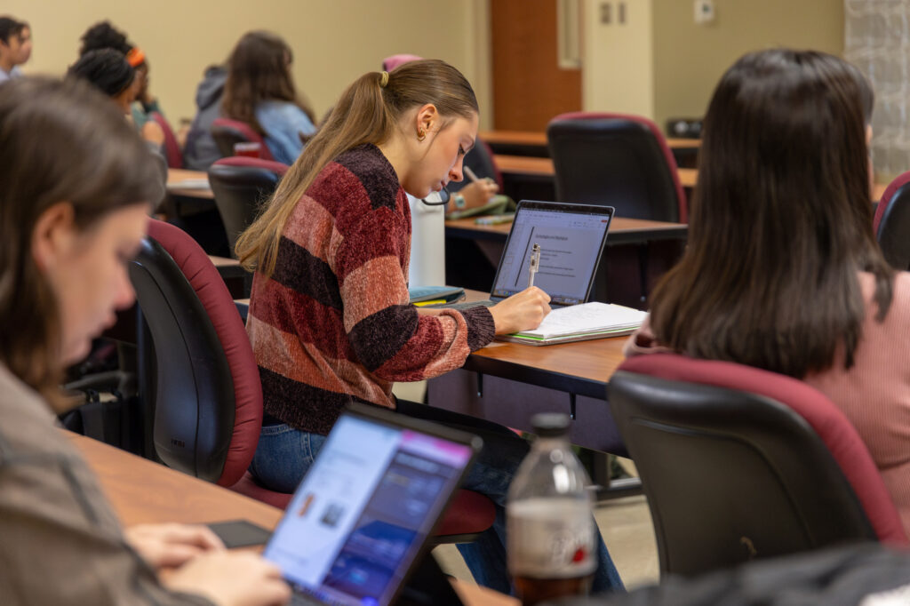 Students sit at desks in a classroom with laptops and notebooks. One student in the center writes in a notebook while looking at a laptop screen. Other students focus on their screens. A bottle is on one desk.