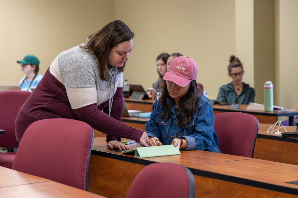 A woman stands next to a seated student, pointing at a tablet on a desk in a classroom. Other students sit at desks in the background, some using laptops.