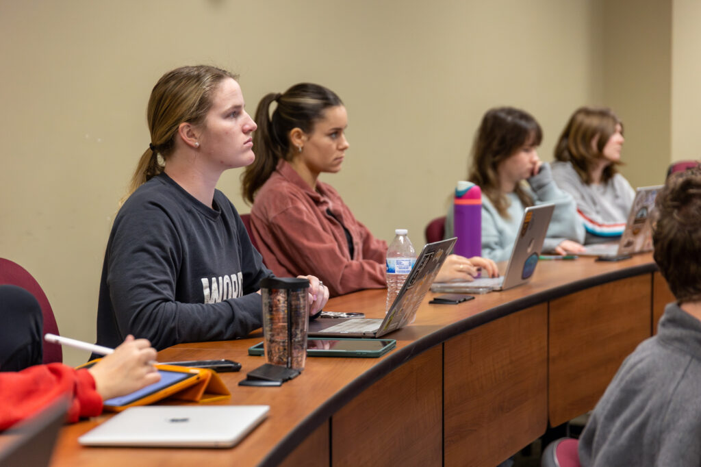 Students sit at curved desks in a classroom, focused on something out of view. They have laptops, notebooks, and water bottles in front of them. The background is plain, and everyone appears attentive.