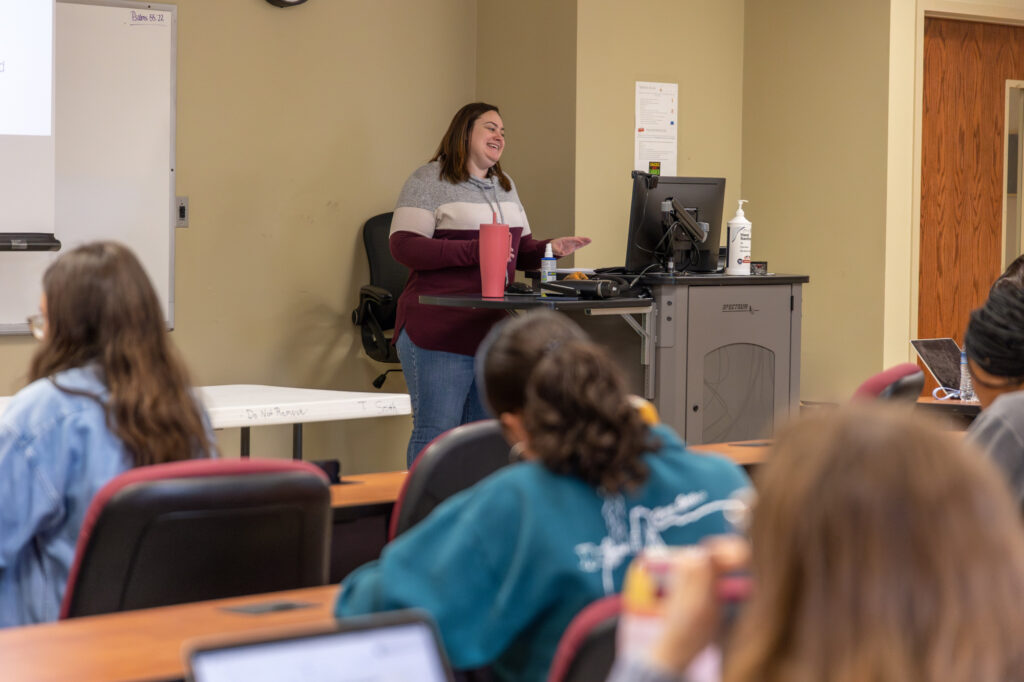 A woman stands at a lectern, speaking to a classroom of seated students. There is a computer monitor and a bottle of sanitizer on the desk near her, and students are taking notes or using laptops.