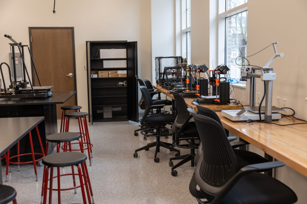 A makerspace featuring 3D printers on a wooden counter, black chairs, red stools at a black table, shelves, and a closed door. Large windows let in natural light. The space is tidy and organized.