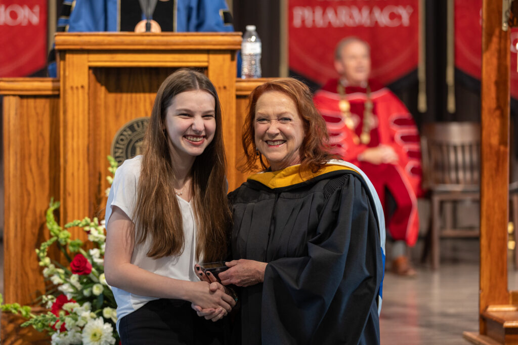 A young woman smiles while receiving an award from a faculty member in academic regalia during a ceremony. A podium, red banners, and another person seated in academic dress are visible in the background.