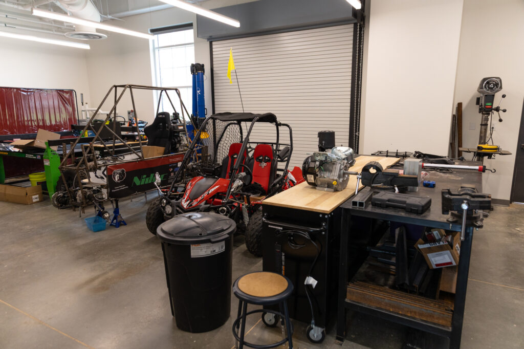 A workshop with a red off-road go-kart, a metal frame vehicle, workbenches, tools, a trash can, and a stool. There is a large garage door at the back and various equipment and supplies around the room.