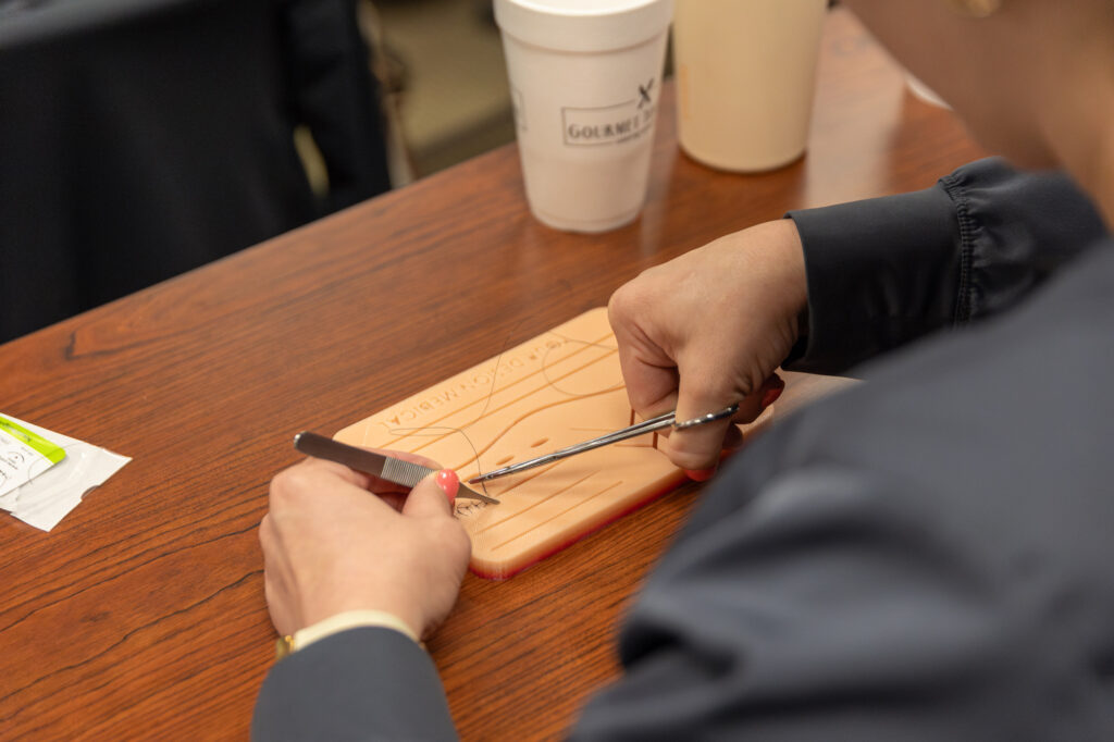 A person practices suturing on a synthetic skin pad with a needle holder and forceps. There are disposable cups and other medical training supplies on the wooden table.