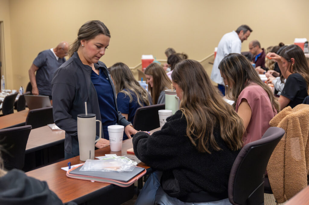 A classroom with several people sitting at desks. A woman stands next to a seated student, both focused on materials in front of them. Other students are working or talking in the background.