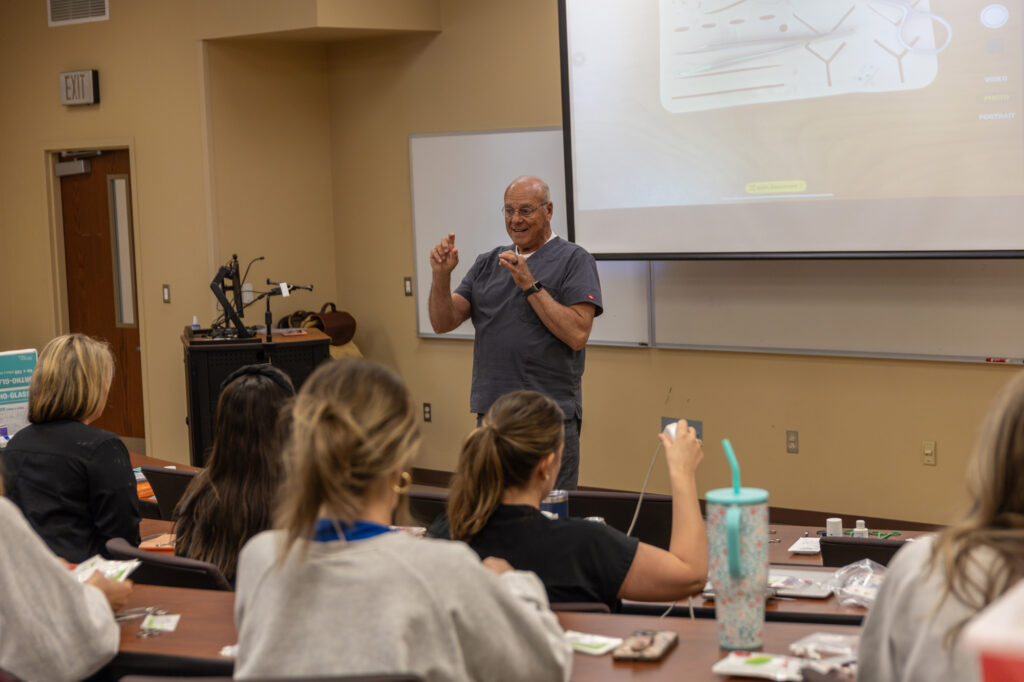 A man stands at the front of a classroom giving a lecture to students seated at desks. A large screen with diagrams is displayed behind him, and various items are spread out on the students desks.