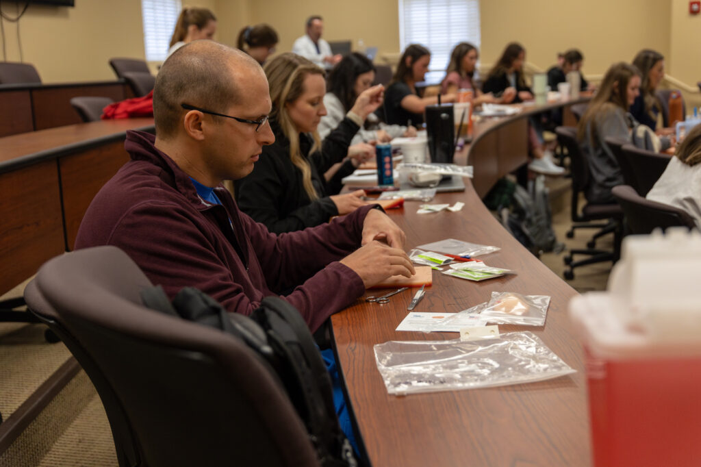 Several people sit at desks in a classroom, handling medical supplies and practice materials, possibly participating in a hands-on medical or nursing skills workshop. The focus is on a man working with tools at the front.