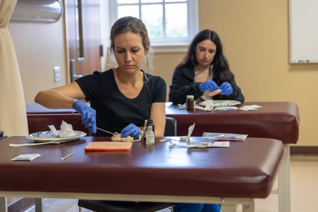 Two women in gloves sit at separate tables in a classroom, working on medical practice materials with tools and supplies in front of them. A window and whiteboard are visible in the background.