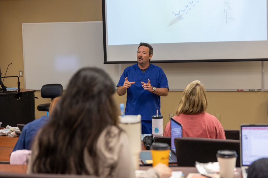 A man in blue scrubs stands at the front of a classroom, gesturing while speaking to seated students. A projection screen behind him displays a diagram. Laptops, notebooks, and drinks are visible on students desks.