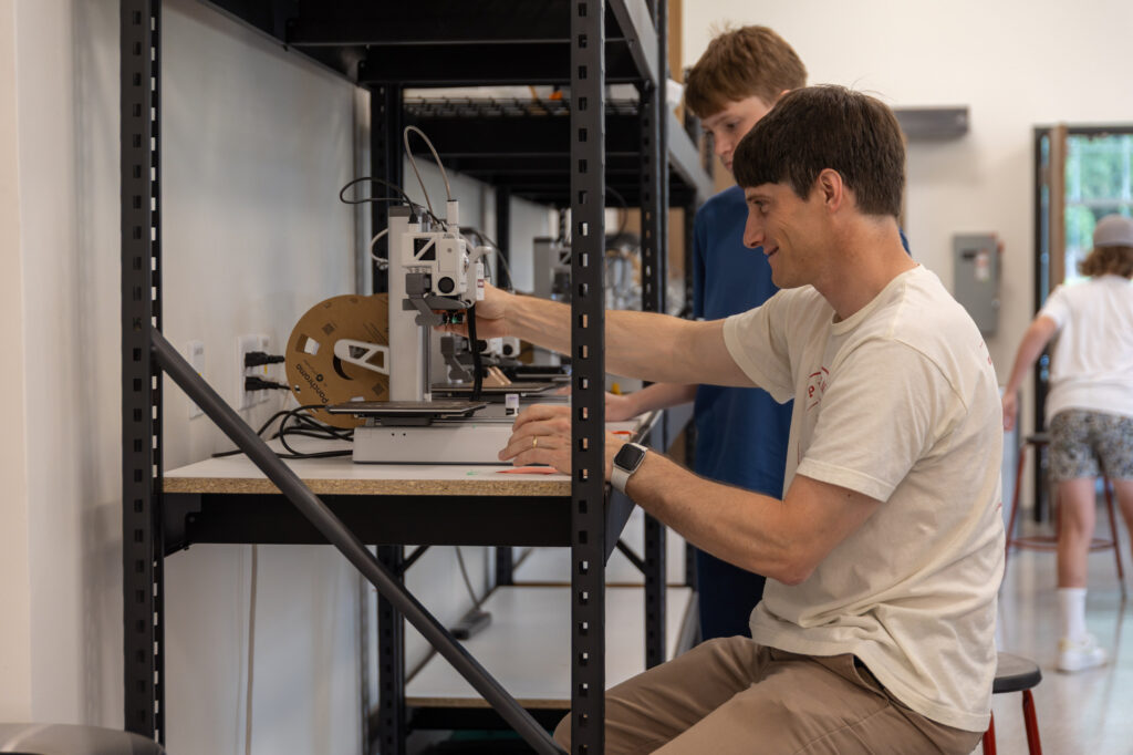 A man in a white shirt operates a 3D printer on a workbench in a lab. Another person stands behind him, and a third person is in the background near a doorway. There are shelves and various equipment in the room.
