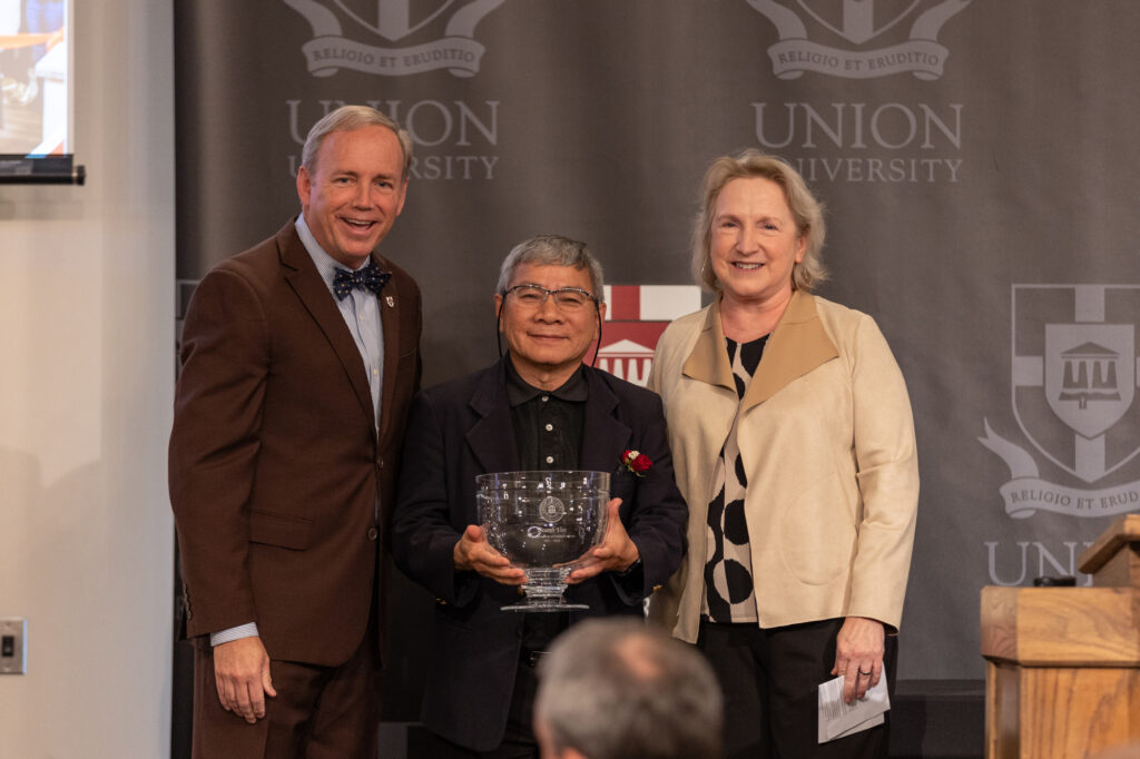 Three people stand in front of a Union University backdrop. The man in the middle holds a large glass award. The man on the left wears a brown suit, and the woman on the right wears a beige jacket. All are smiling.