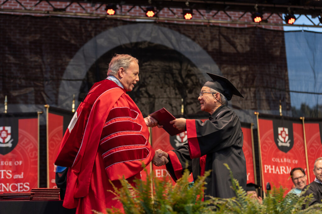A person in academic regalia receives a diploma from another person in a red gown during an outdoor graduation ceremony, with university banners in the background.