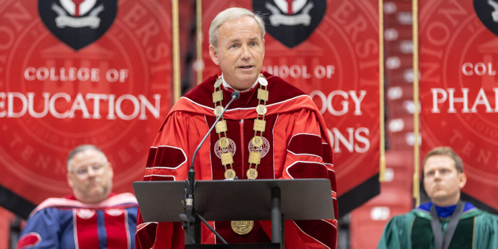 A person in academic regalia stands at a podium speaking at a fall convocation graduation ceremony. Two people in academic robes sit behind him. Banners for the College of Education and School of Pharmacy hang in the background, reflecting on how education prepares graduates to serve Christ in a turning world.