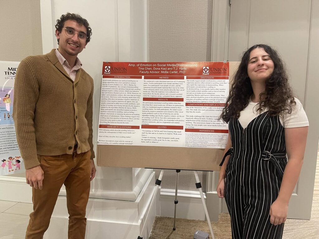 Two people stand beside a research poster titled App. of Emotion on Social Media Platforms at Union College. Both are smiling; one wears a brown sweater and glasses, the other wears a striped dress over a white shirt.