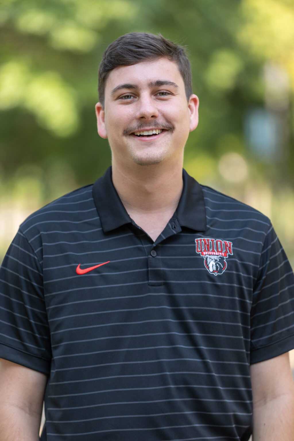A man with short brown hair and a mustache smiles, wearing a black, striped Union Bulldogs polo shirt. He stands outdoors with a blurred background of greenery.