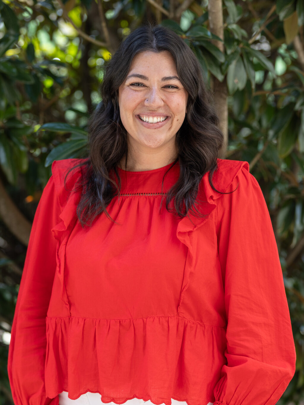 A woman with long dark hair, wearing a bright red ruffled blouse, stands outside in front of leafy green branches, smiling at the camera.