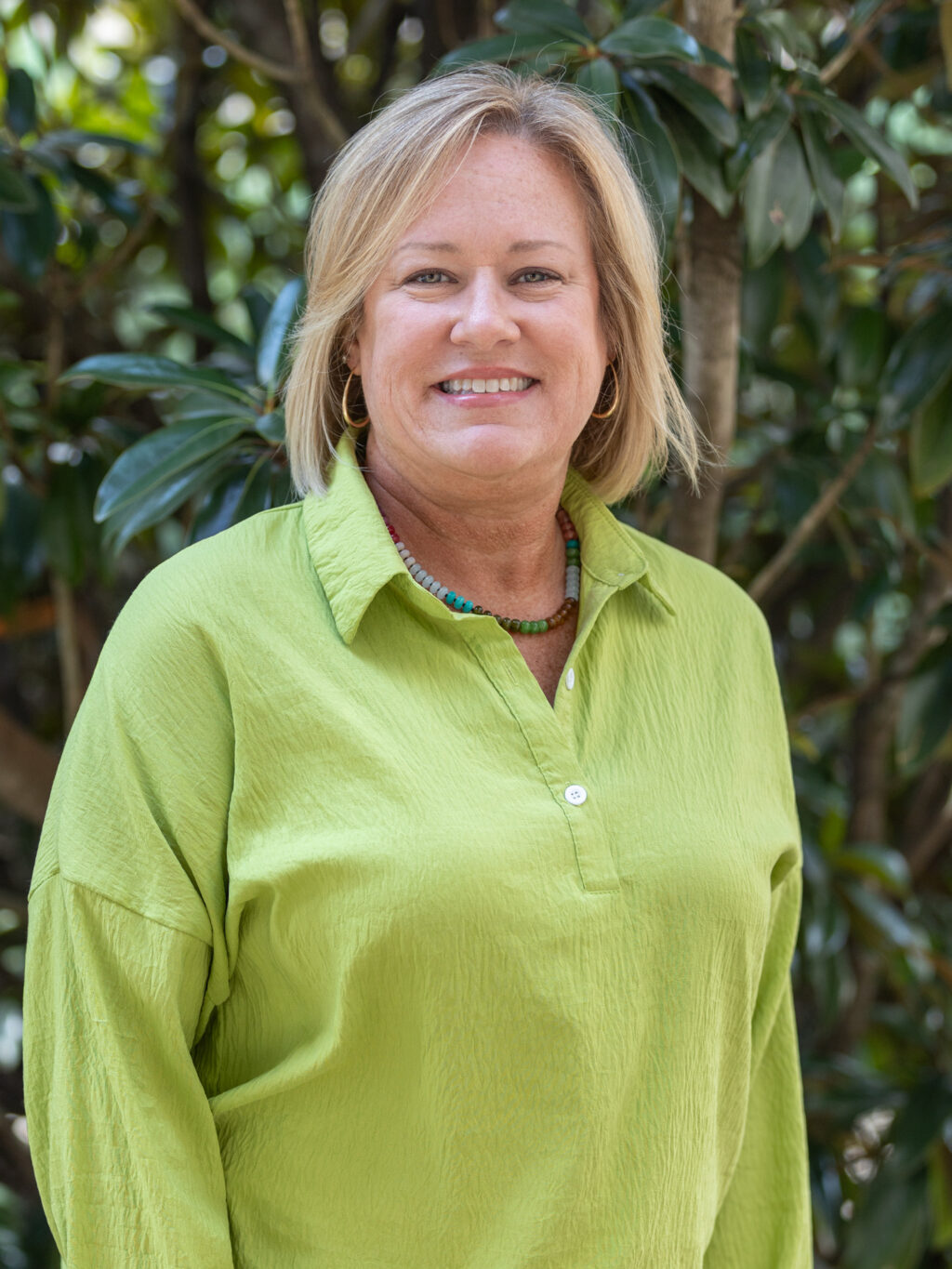 A woman with blond hair wearing a light green shirt and a beaded necklace stands outdoors in front of leafy trees, smiling at the camera.