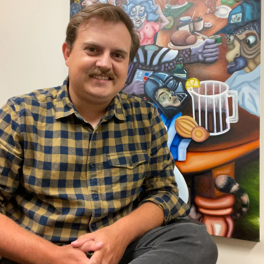 A person with a mustache and checked shirt sits smiling in front of a colorful painting that features cartoonish, animal-like characters around a table with food and drinks, capturing the lively spirit of an art gallery exhibition by Clay Palmer.