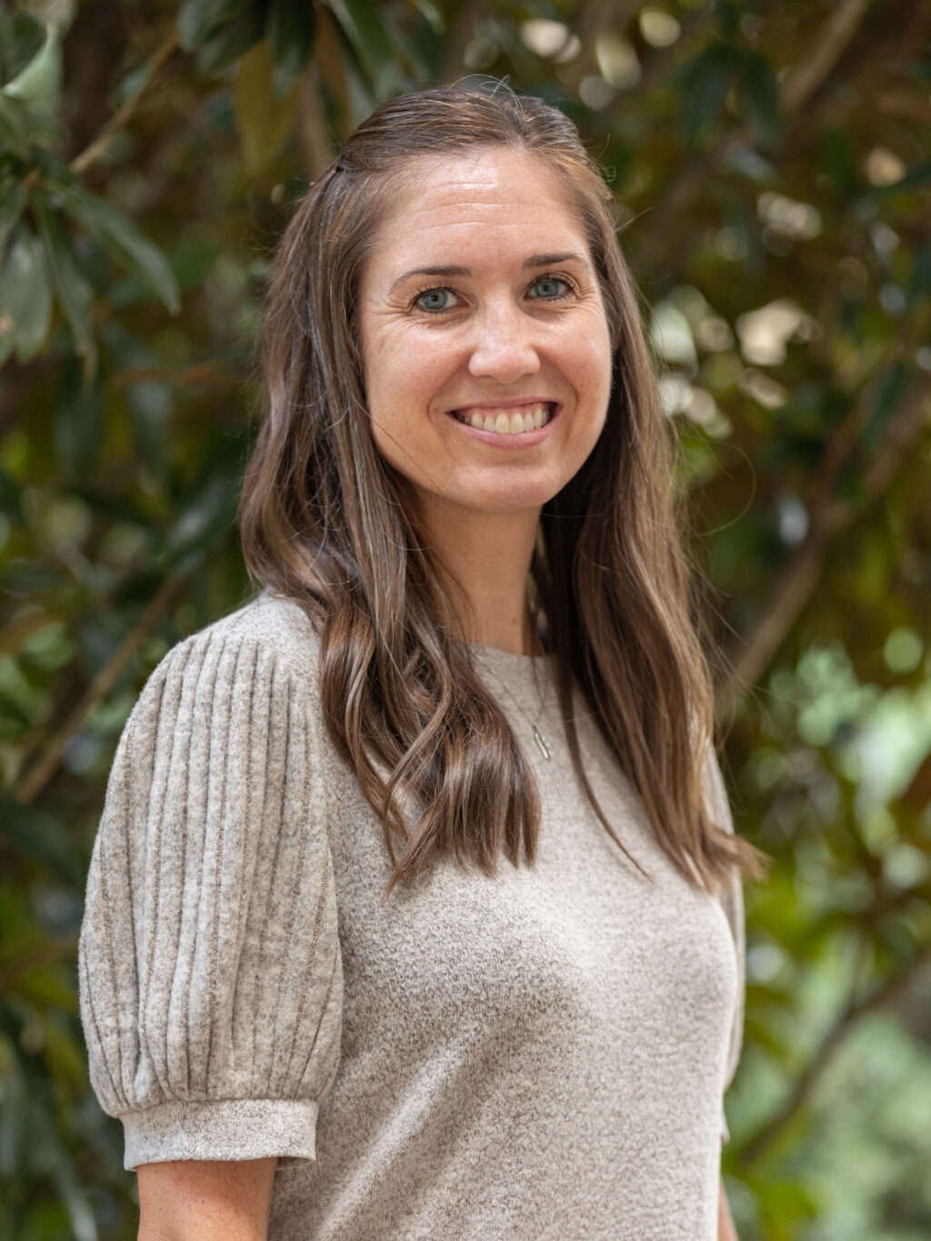 A woman with long brown hair and a light gray short-sleeved sweater stands outdoors in front of green foliage, smiling at the camera.