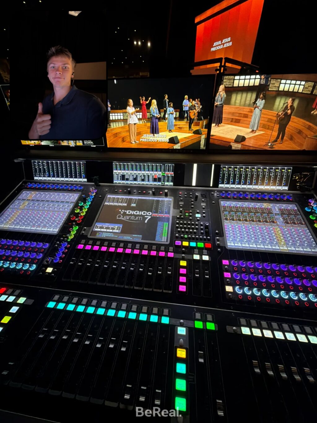 A large audio mixing console with colorful buttons and screens in a control room; video monitors above show people performing on stage, while Jordyn Youngblood gives a thumbs-up gesture.