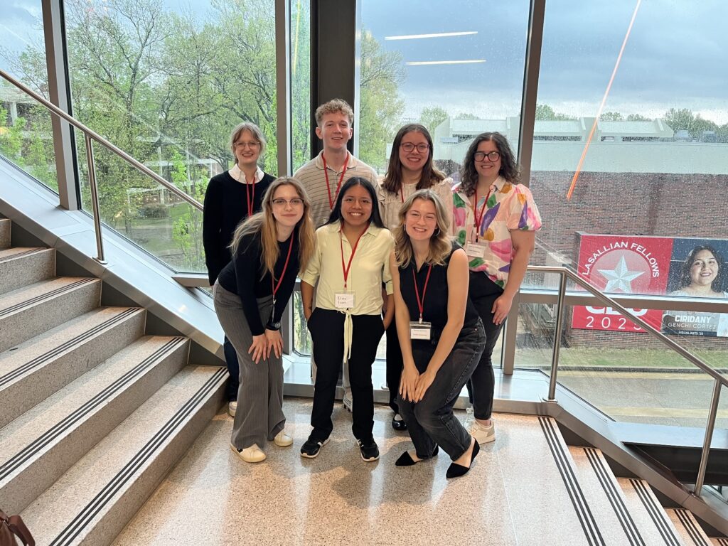 Seven people with name tags and red lanyards pose on indoor stairs in front of large windows. Trees and a building with a red Fellows 2023 banner are visible outside. Some people are standing, others are kneeling.