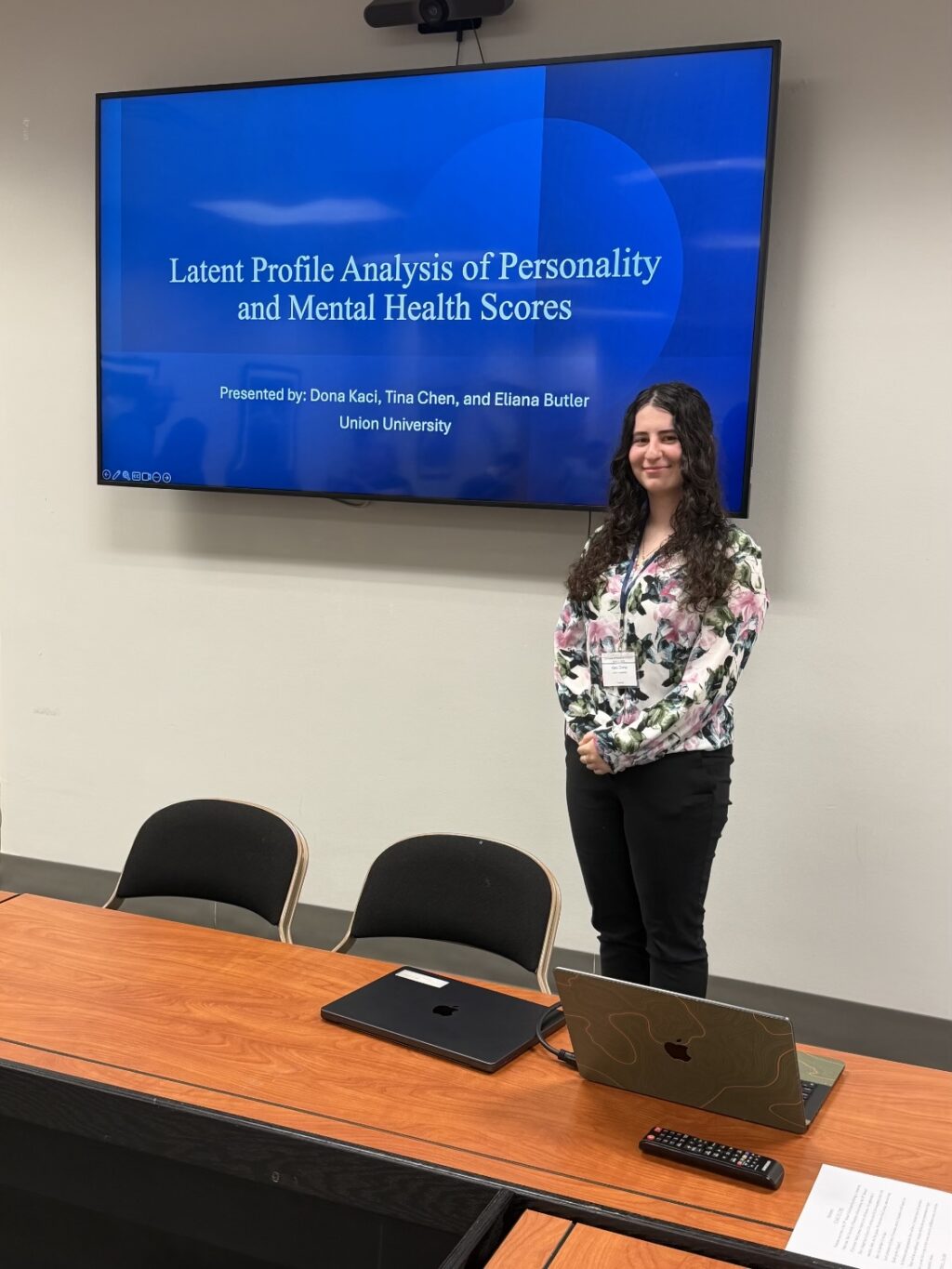A woman stands next to a conference table in front of a screen displaying a presentation titled Latent Profile Analysis of Personality and Mental Health Scores at Union University. A laptop and mouse are on the table.