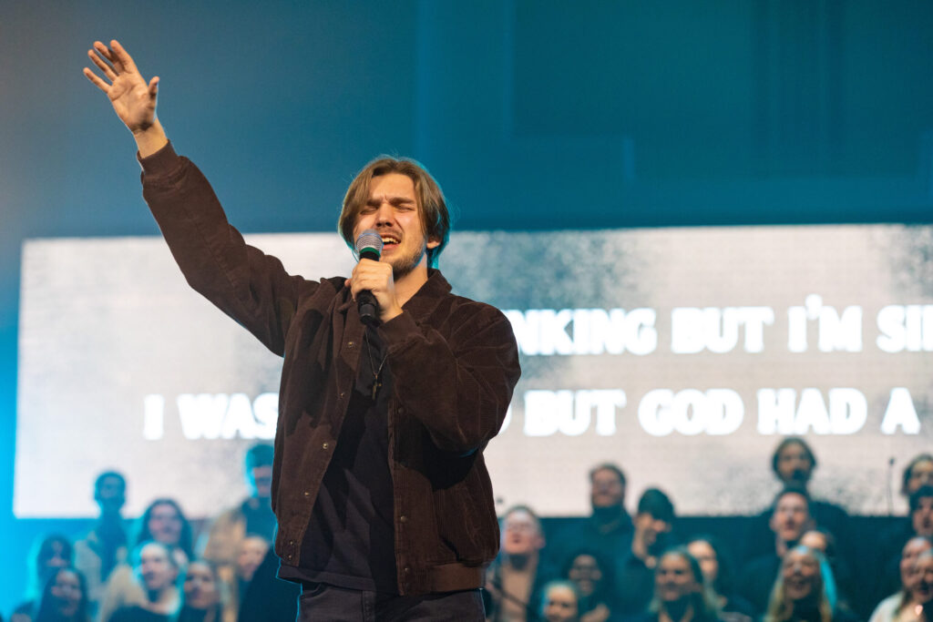 Isaac Reid stands on stage holding a microphone with one hand raised, embodying the lively energy of a Jordyn Youngblood performance. He appears to be singing while a blurred group of people and a large screen with partially readable text form the backdrop. Blue and yellow lights illuminate the scene, adding vibrancy to the moment.