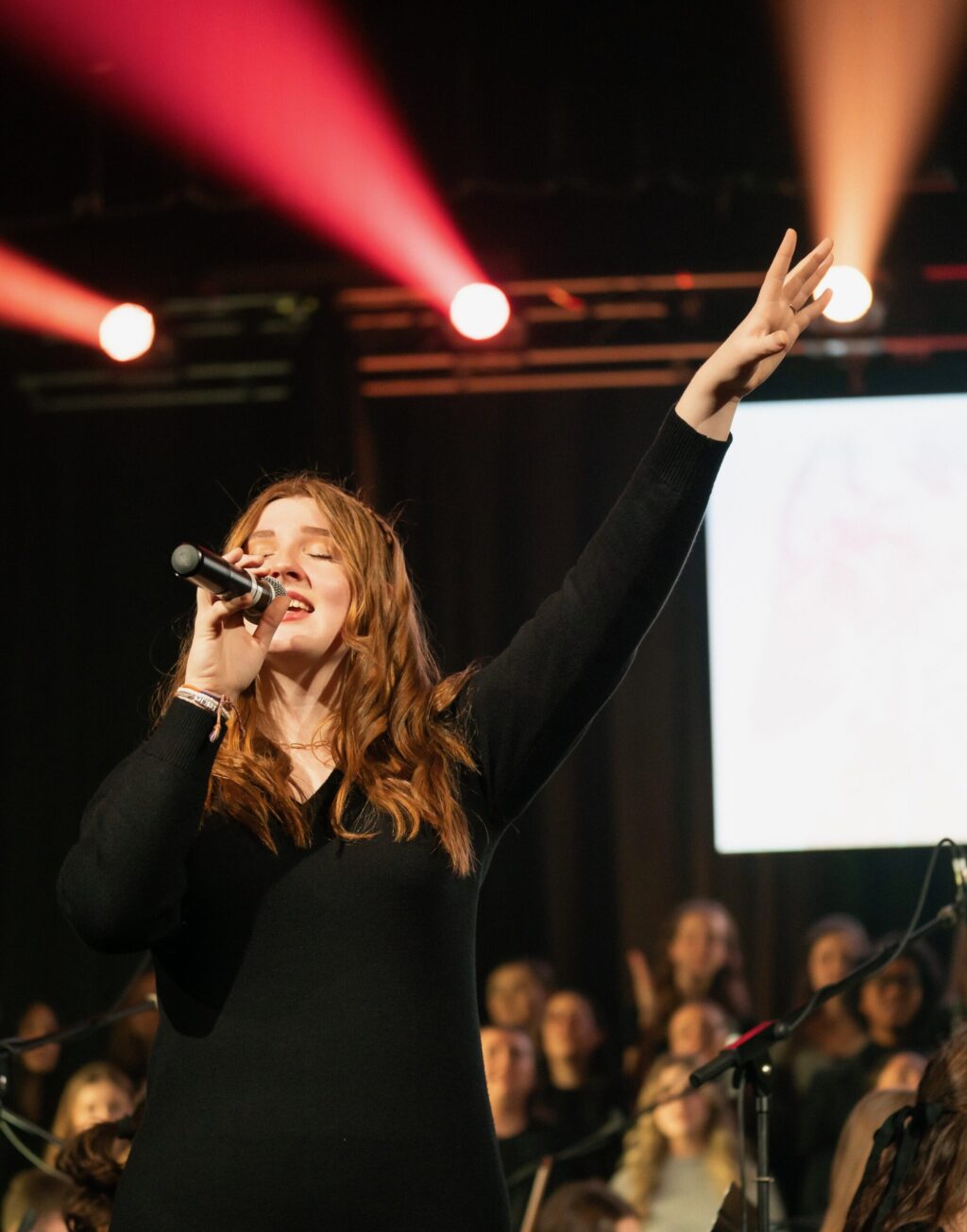 Jordyn Youngblood, dressed in a black dress, sings into a microphone on stage, raising one hand. Red stage lights shine in the background while an audience and other performers are visible behind her.