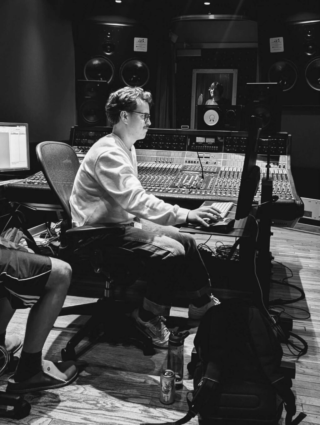 Jordyn Youngblood sits at a large mixing console in a recording studio, focused intently on using the computer. Another person’s legs are partially visible beside them, while studio equipment and speakers surround the scene. The photo is captured in striking black and white.