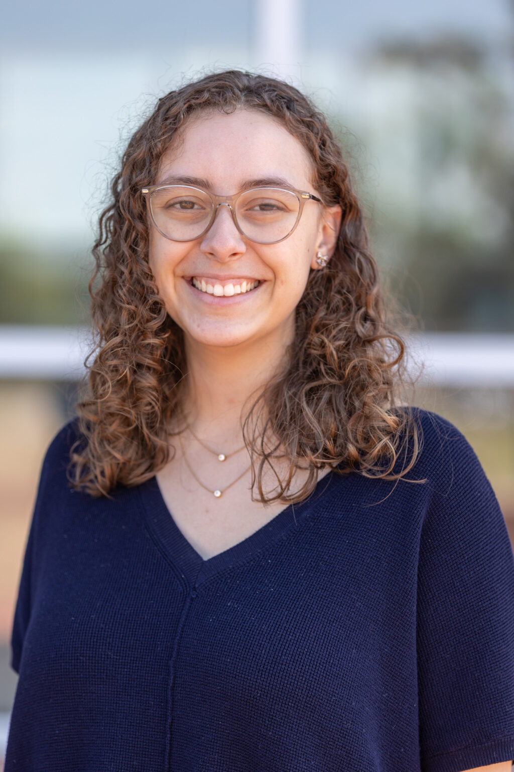 A person with curly brown hair and glasses is smiling at the camera. They are wearing a dark blue top and a delicate necklace. The background is softly blurred, showing some greenery and a window.