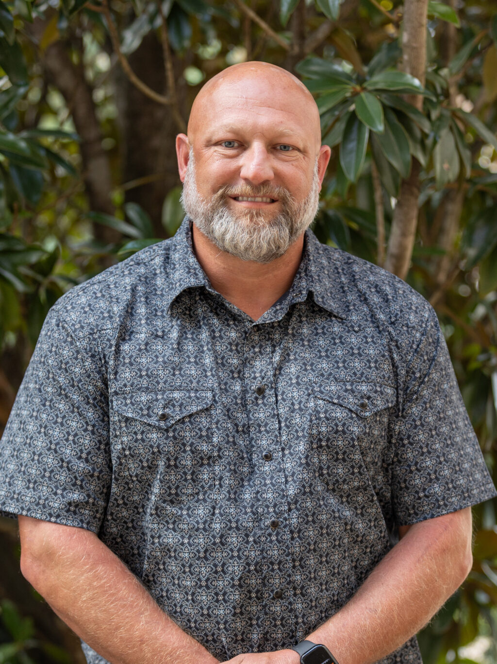 A bald man with a gray beard wearing a short-sleeve patterned button-up shirt stands outdoors in front of green foliage, smiling at the camera with his hands clasped in front of him.