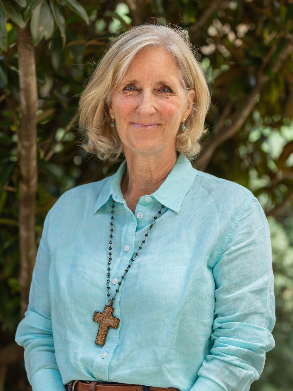A woman with short gray hair stands outdoors in front of leafy trees. She is wearing a light blue button-up shirt, a brown belt, and a wooden cross necklace, and she smiles at the camera.