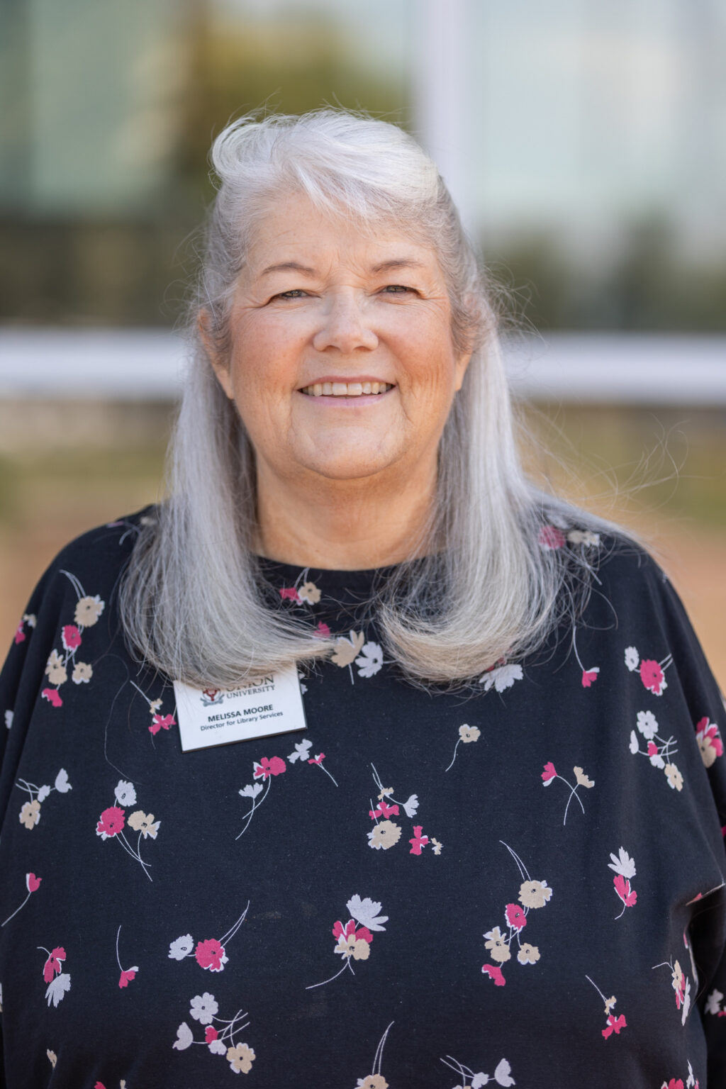 An older woman with long gray hair and a floral top stands outdoors, smiling. She is wearing a name tag. The background is blurred.