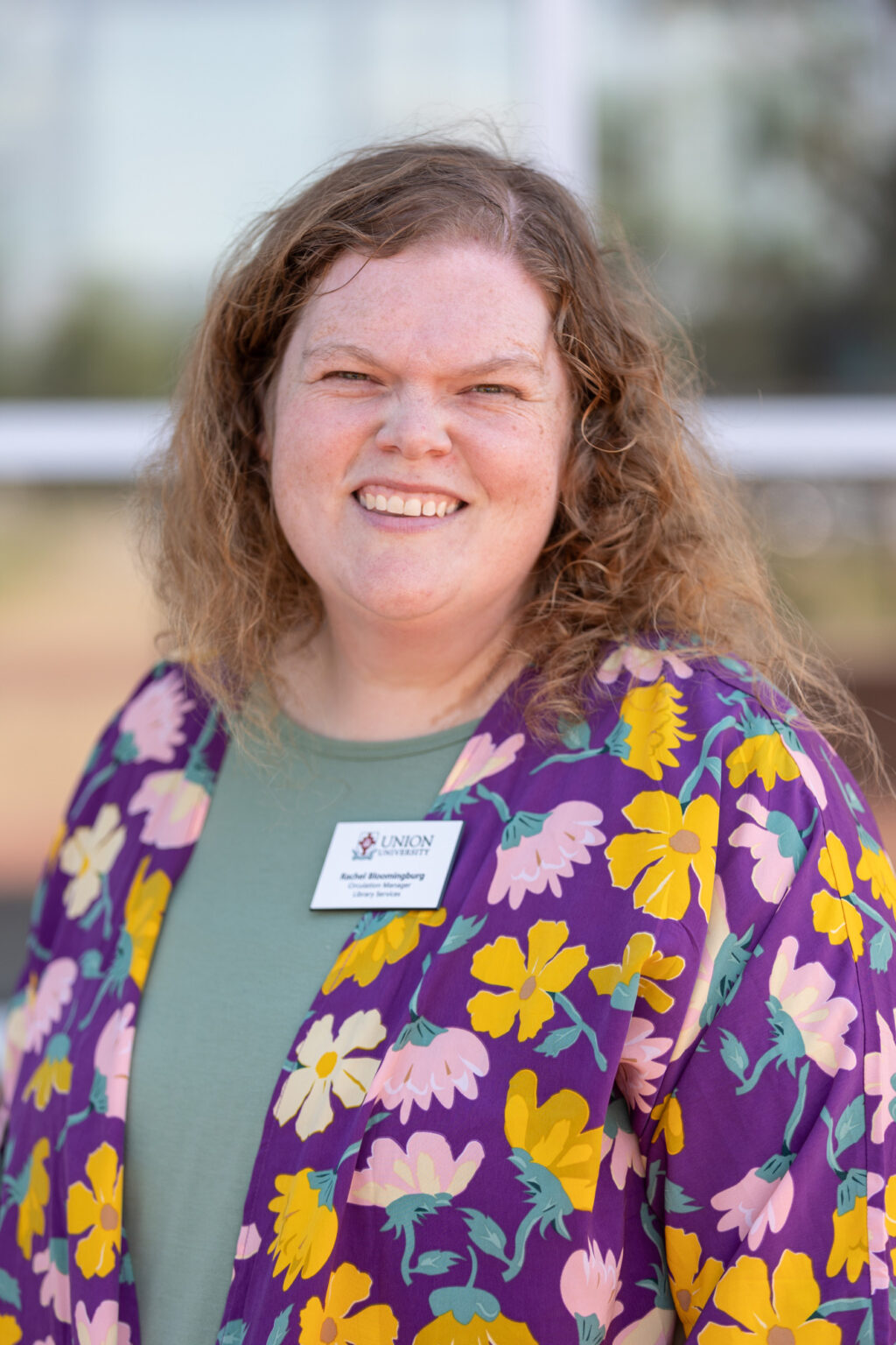 A person with curly hair smiles at the camera, wearing a floral purple and yellow cardigan over a green shirt. They have a Union Public Schools name badge. The background is blurred with some greenery and a window visible.