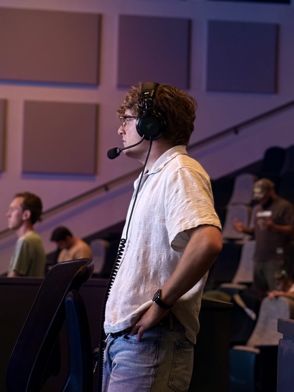 Ryan Cipkowski, wearing a headset and light shirt, stands with one hand in their pocket, looking forward. Other people sit and stand in the background of a room with acoustic panels on the wall.