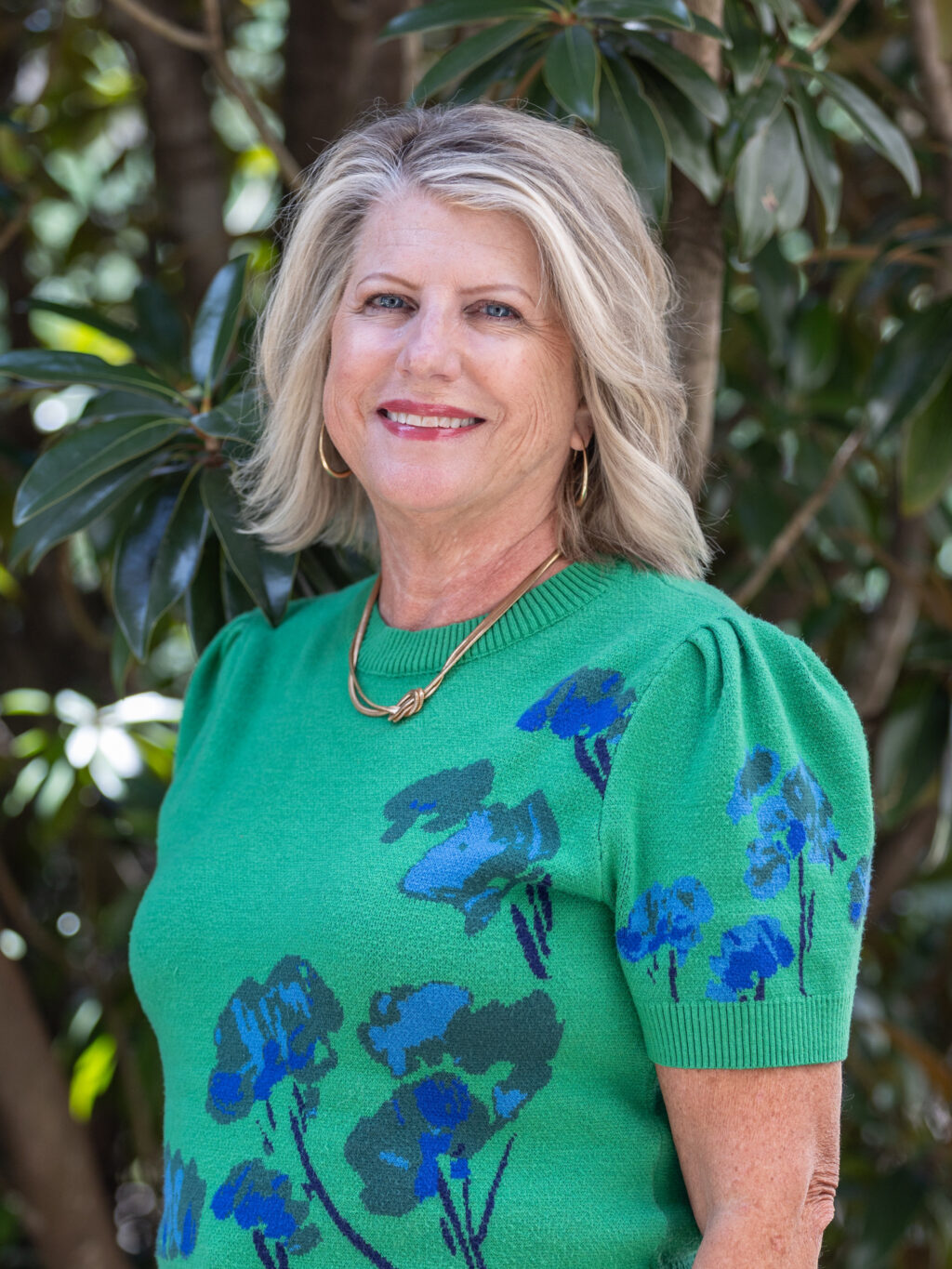 A woman with shoulder-length blonde hair is standing outdoors in front of green foliage, wearing a green sweater with blue flower prints and a gold necklace. She is smiling and looking at the camera.