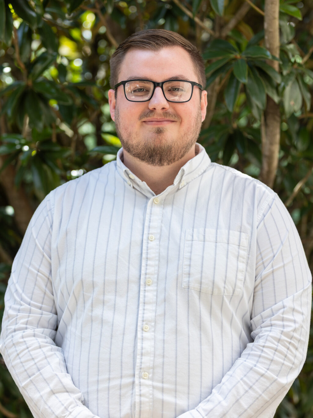 A man with short brown hair, a beard, and glasses wears a white, long-sleeved, striped button-up shirt. He stands outdoors in front of leafy green trees, looking at the camera with a neutral expression.