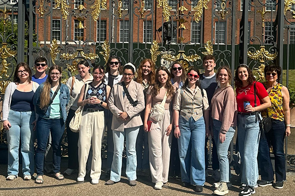 A group of 16 people stands together in front of an ornate black and gold gate on a sunny day in the UK. Most are smiling and wearing casual clothing, with some holding sunglasses or water bottles. Brick buildings are visible in the background.