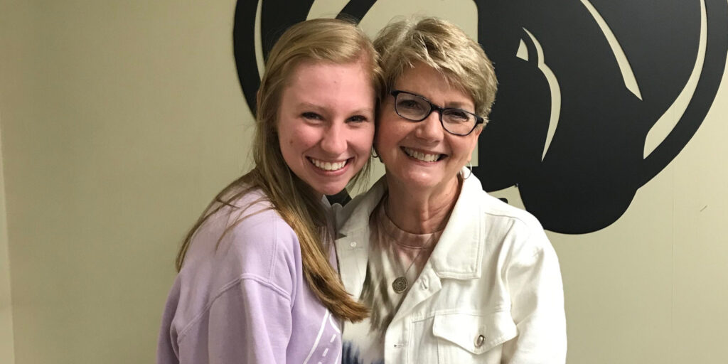 Two women, one younger and one older, are standing close together and smiling at the camera. The older woman, Susan Hopper, is wearing glasses and a white jacket. They are in front of a beige wall with a large black graphic, capturing a moment that truly feels like a family’s answer to prayer.