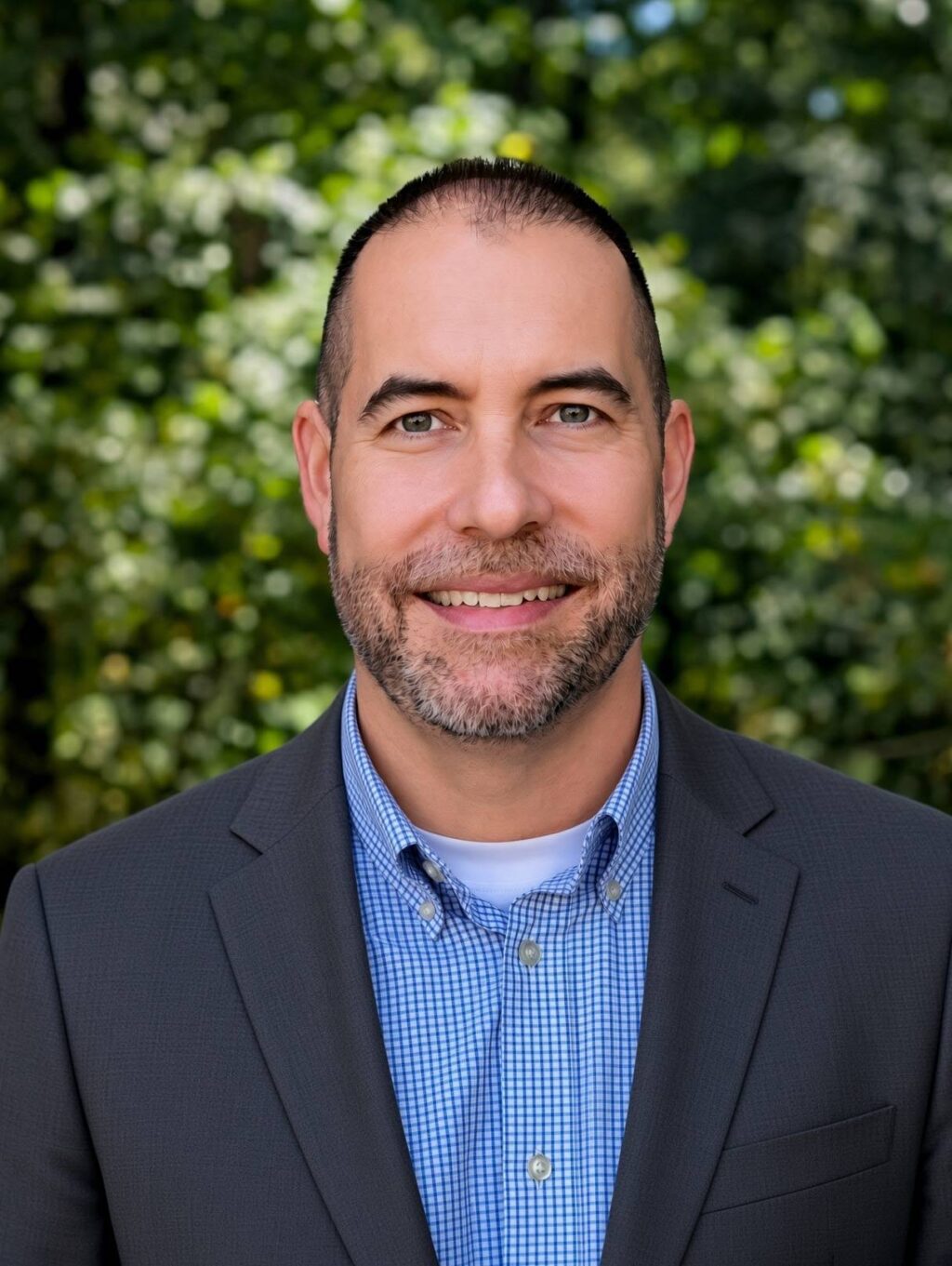 A man with short dark hair and a trimmed beard is wearing a gray suit jacket over a blue checked shirt and white undershirt, standing outdoors in front of green, leafy trees.