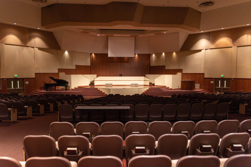Rows of empty seats face a stage in an auditorium with wooden walls. A grand piano is on the left, and a podium is on the right. Overhead lights illuminate the stage, and exit signs are visible on both sides.