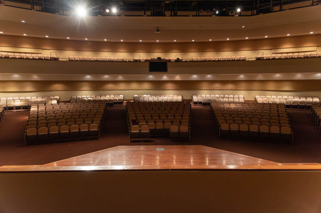View from a wooden stage looking out at an empty auditorium with rows of tan cushioned seats and two balconies, under warm lighting.