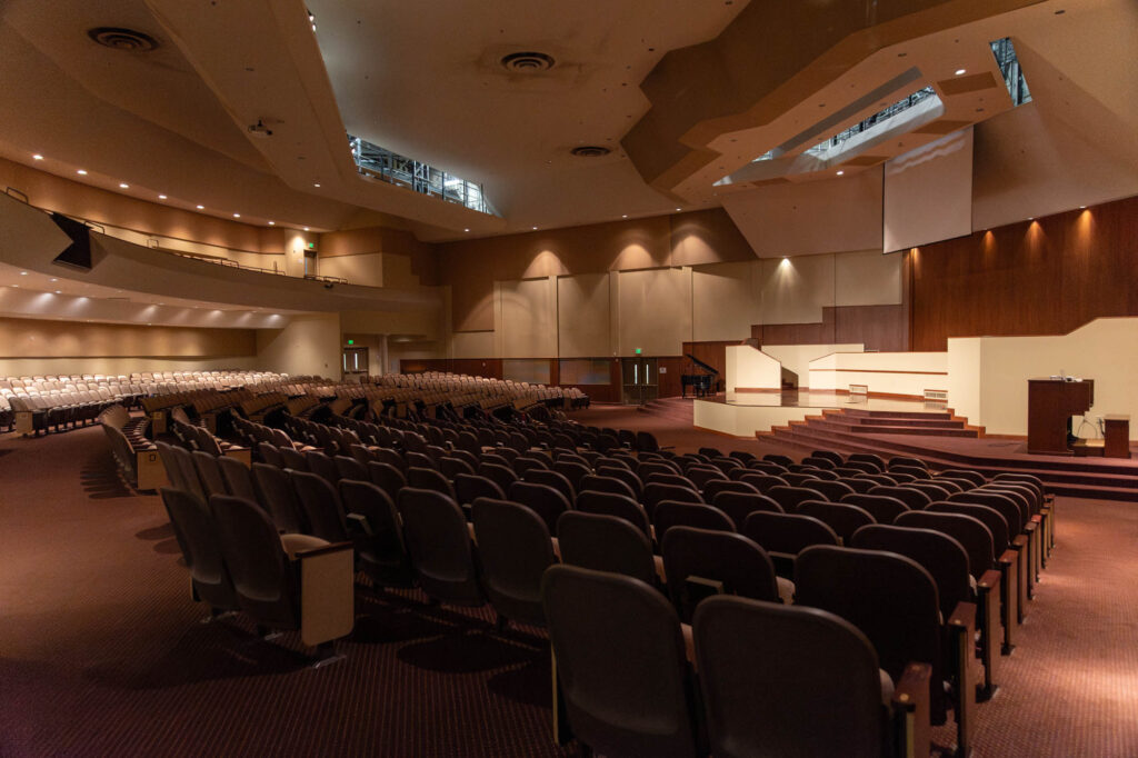 Large, empty auditorium with rows of dark chairs facing a stage. The stage has steps, a podium, and a large wall behind it. Soft lighting illuminates the room, and the ceiling has modern architectural features.